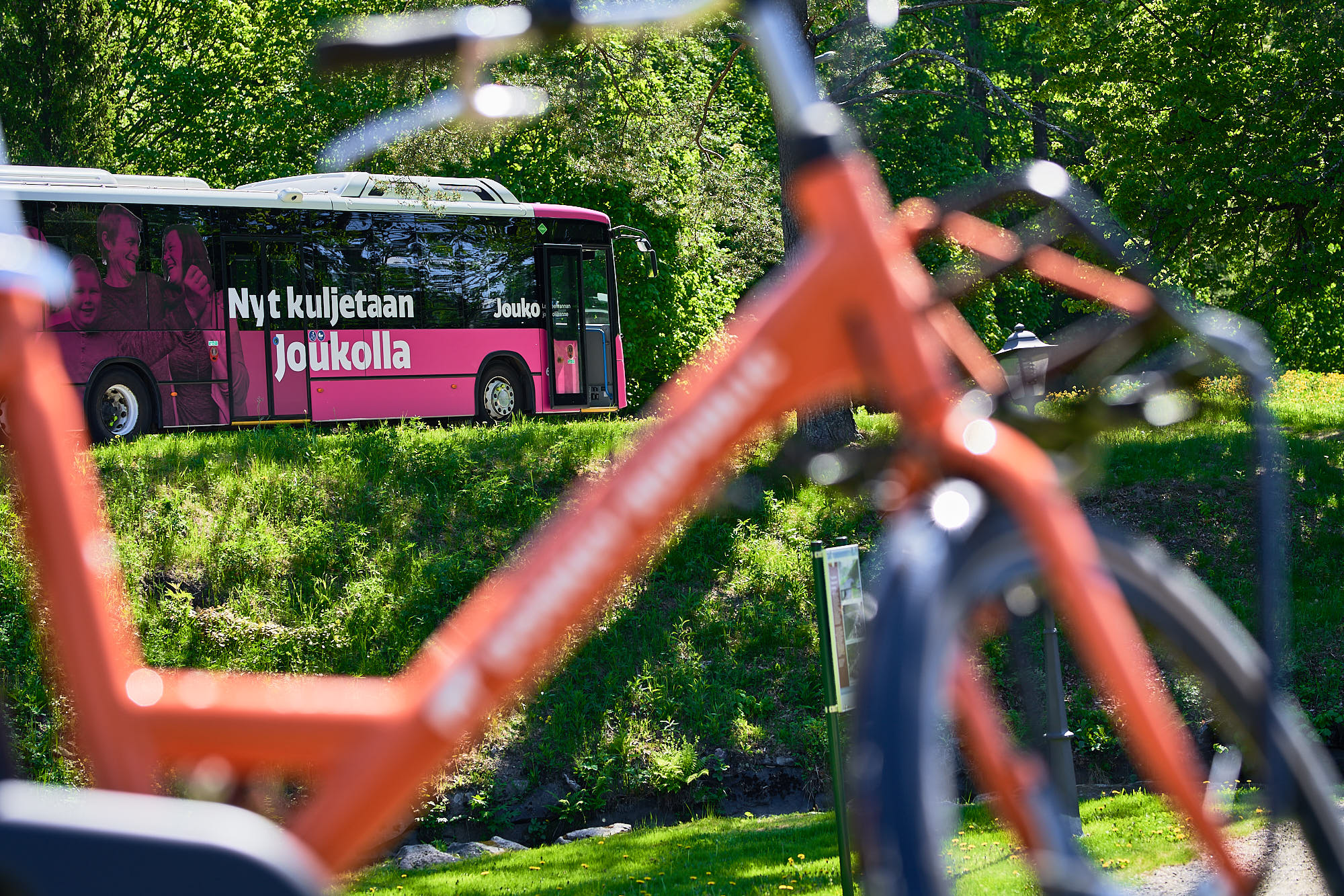 Etualalla oranssi pyörä, taustalla pinkki Jouko-bussi, jossa lukee ”Nyt kuljetaan Joukolla”.