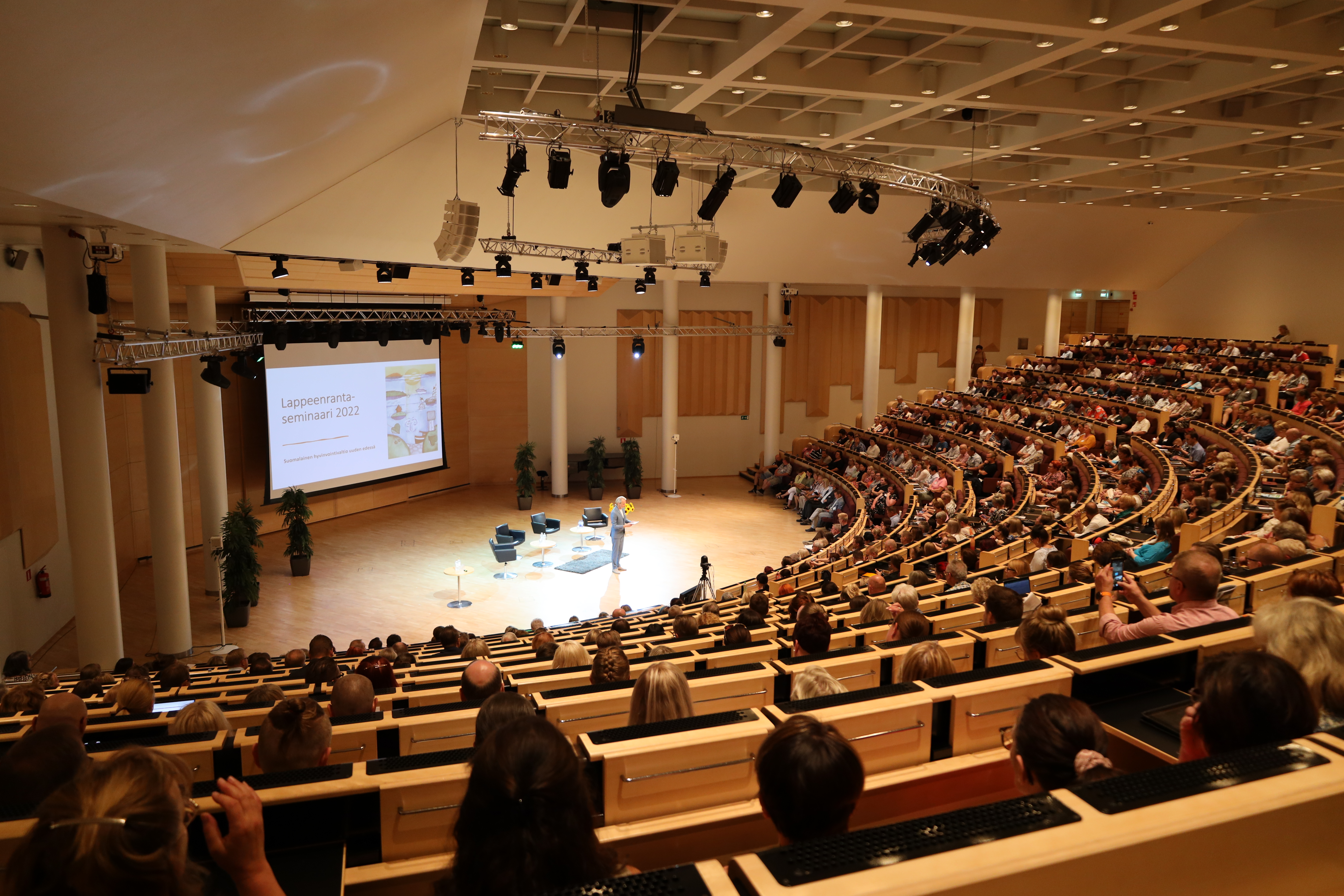 Kuva Lappeenranta-salista, jossa on käynnissä Lappeenranta-seminaari. Ihmiset istuvat katsomon penkeillä ja katsovat lavalla esiintyvää miestä. 