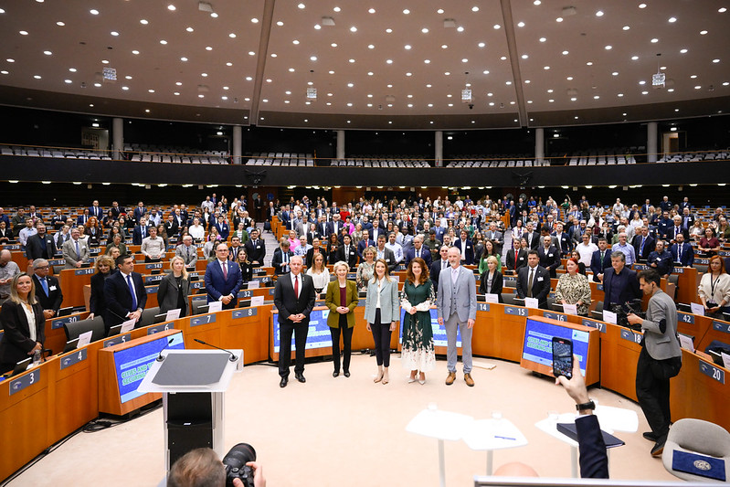 Joukko ihmisistä parlamenttisalissa hymyilee kameraan päin.