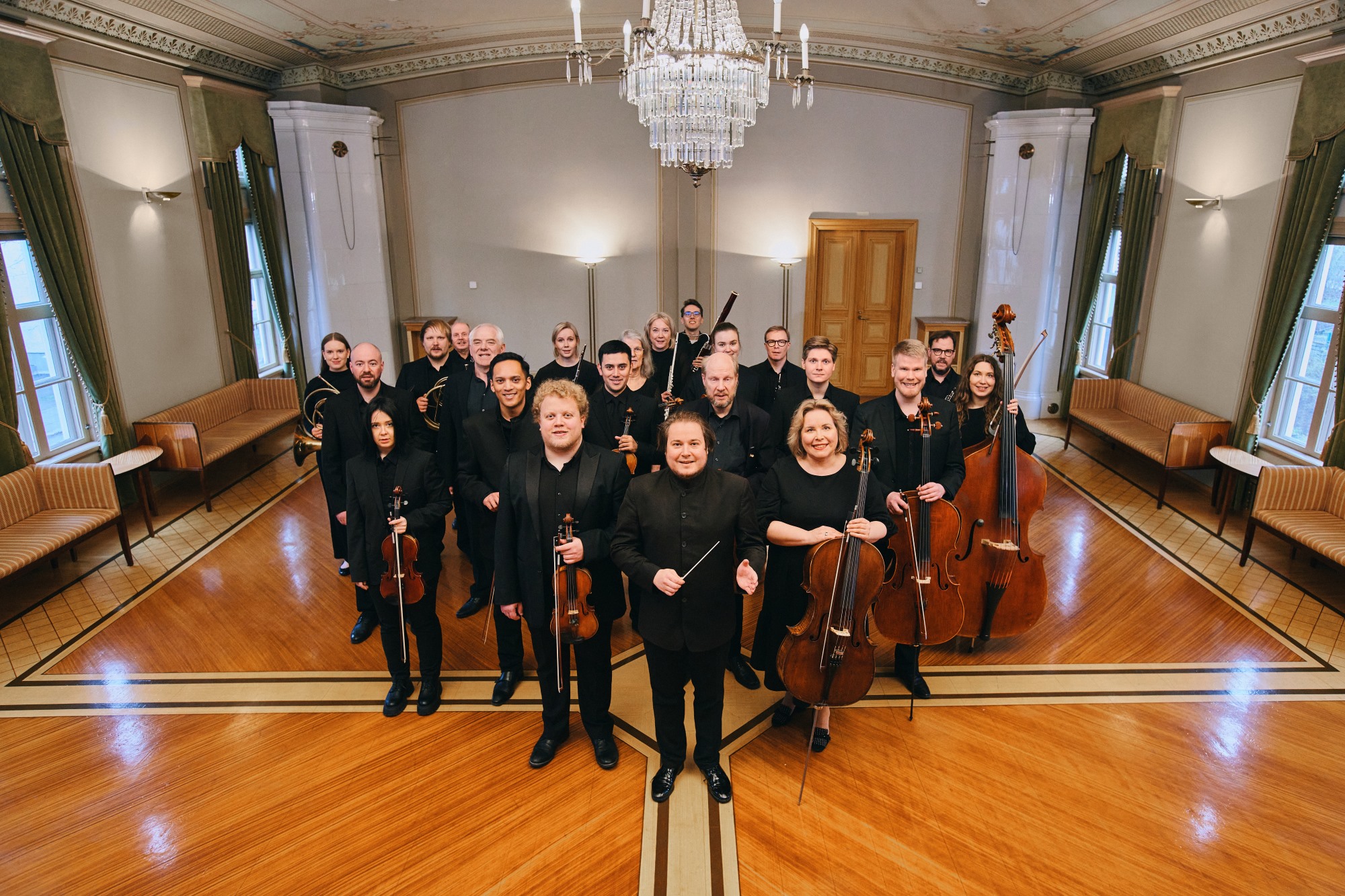 Lappeenrannan kaupunginorkesterin muusikoiden ryhmäkuva. Muusikot seisovat juhlavassa salissa mustiin pukeutuneina ja soittimet kädessä.