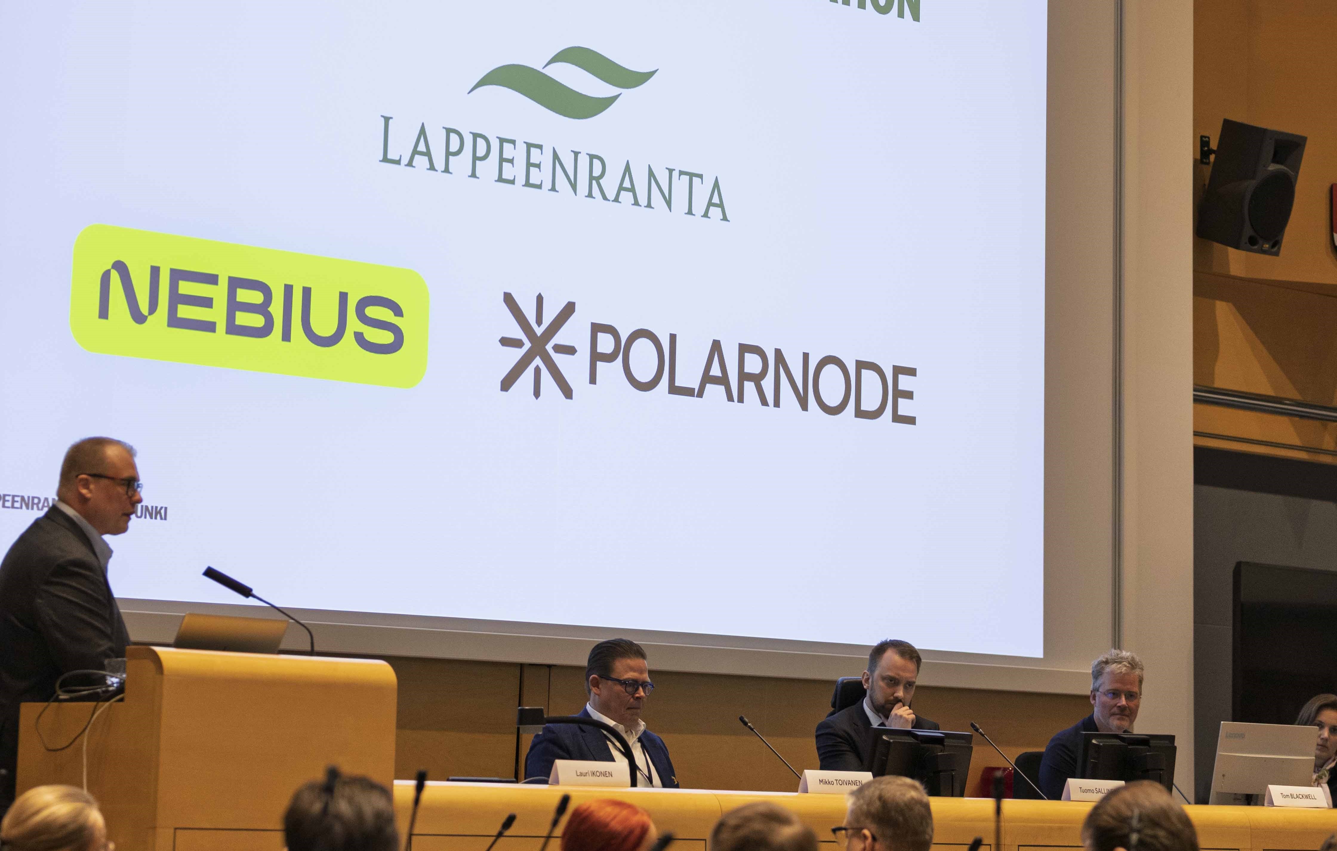 Ihmiset mediatilaisuudessa valtuustosalissa. Näyttöruutu, jossa lukee Yhteistyössä - In collaboration Lappeenranta, Nebius, Polarnode.