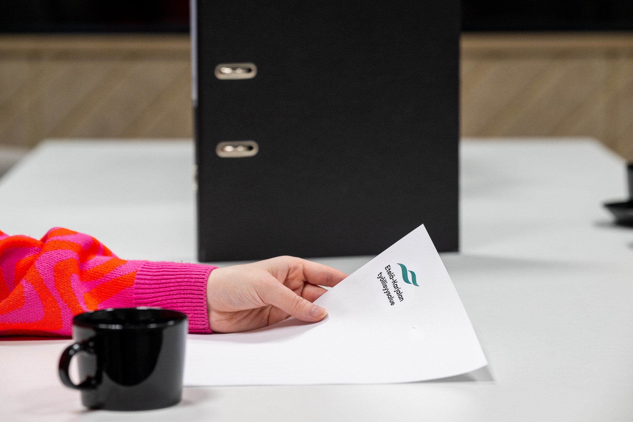 Käsi pitelee paperia, jossa Etelä-Karjalan työllisyysalueen logo. Taustalla mappi, etualalla kahvikuppi.