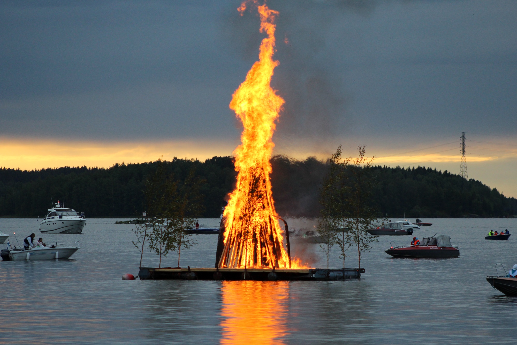 Juhannuskokko palaa Myllysaaren edustalla kesäisenä päivänä. Ihmiset ovat katsomassa kokkoa. 