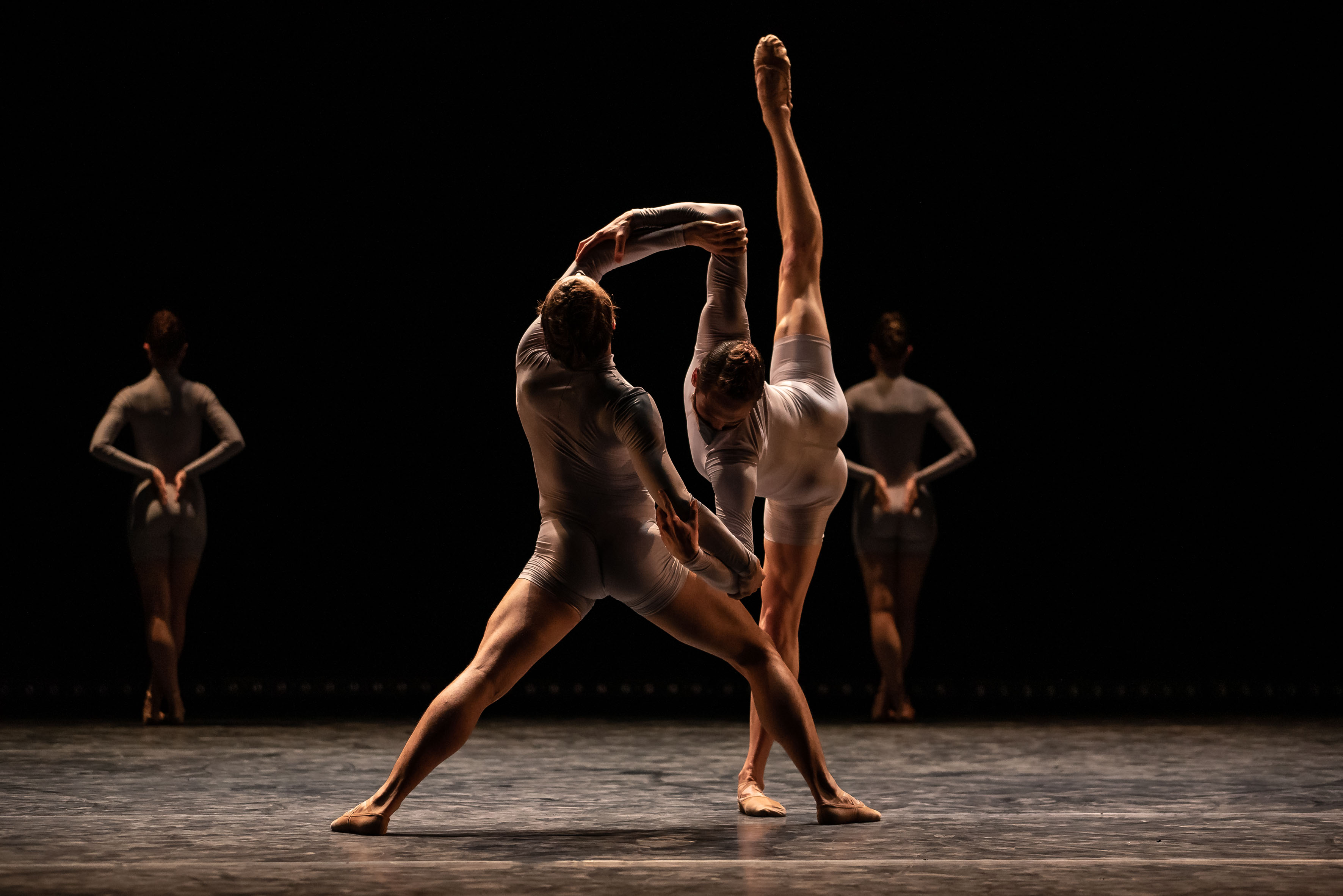 Etualalla kaksi balettitanssijaa tanssimassa dramaattisessa, hämärässä valaistuksessa. Taustalla näkyy kahden muun tanssijan hahmot.