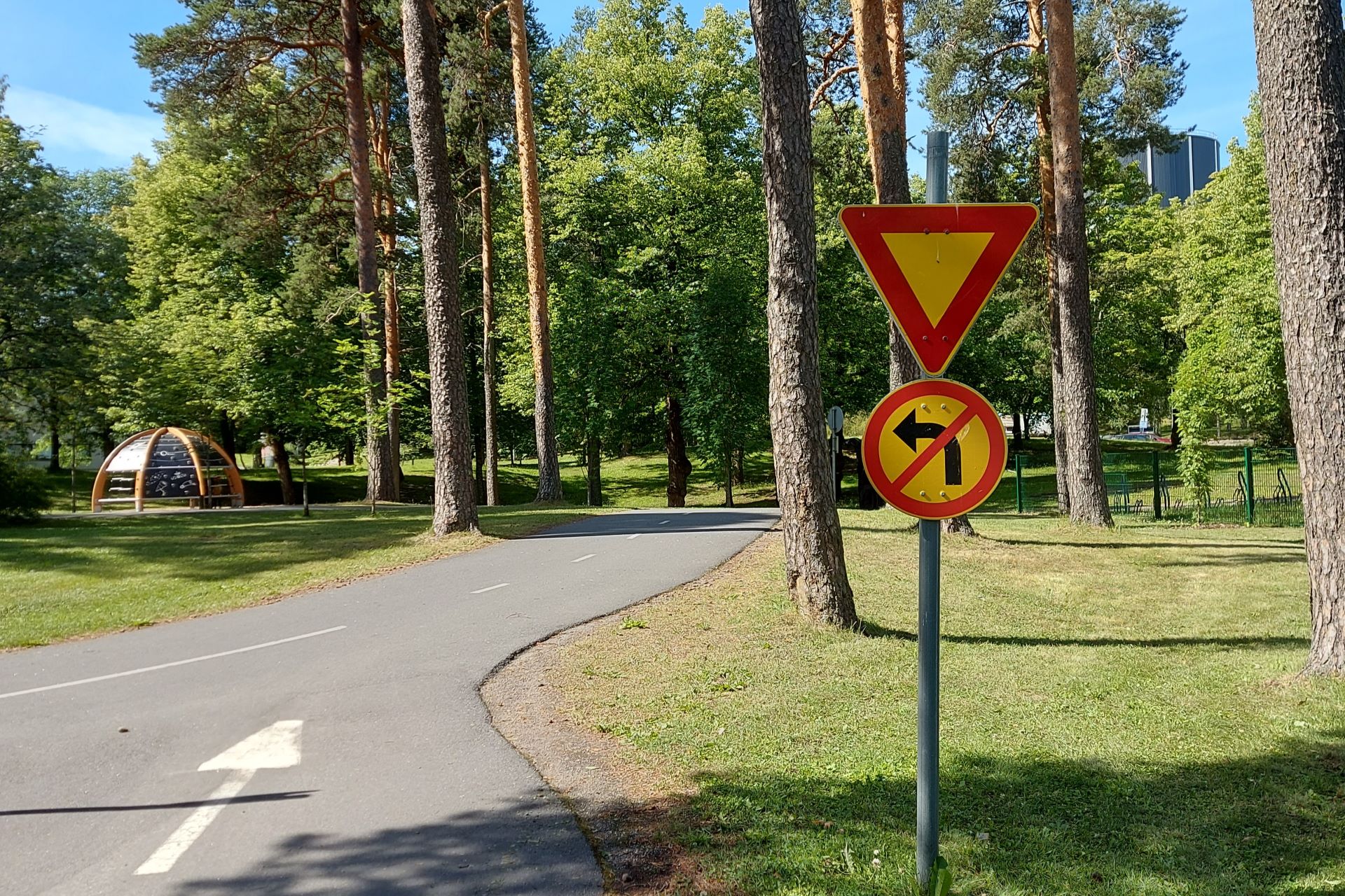 Lasten leikkipuiston ajotie, jonka reunassa on liikennemerkki: väistämisvelvollisuus risteyksessä ja vasemmalle kääntyminen kielletty. Taustalla puustoa ja kiipeilyteline.