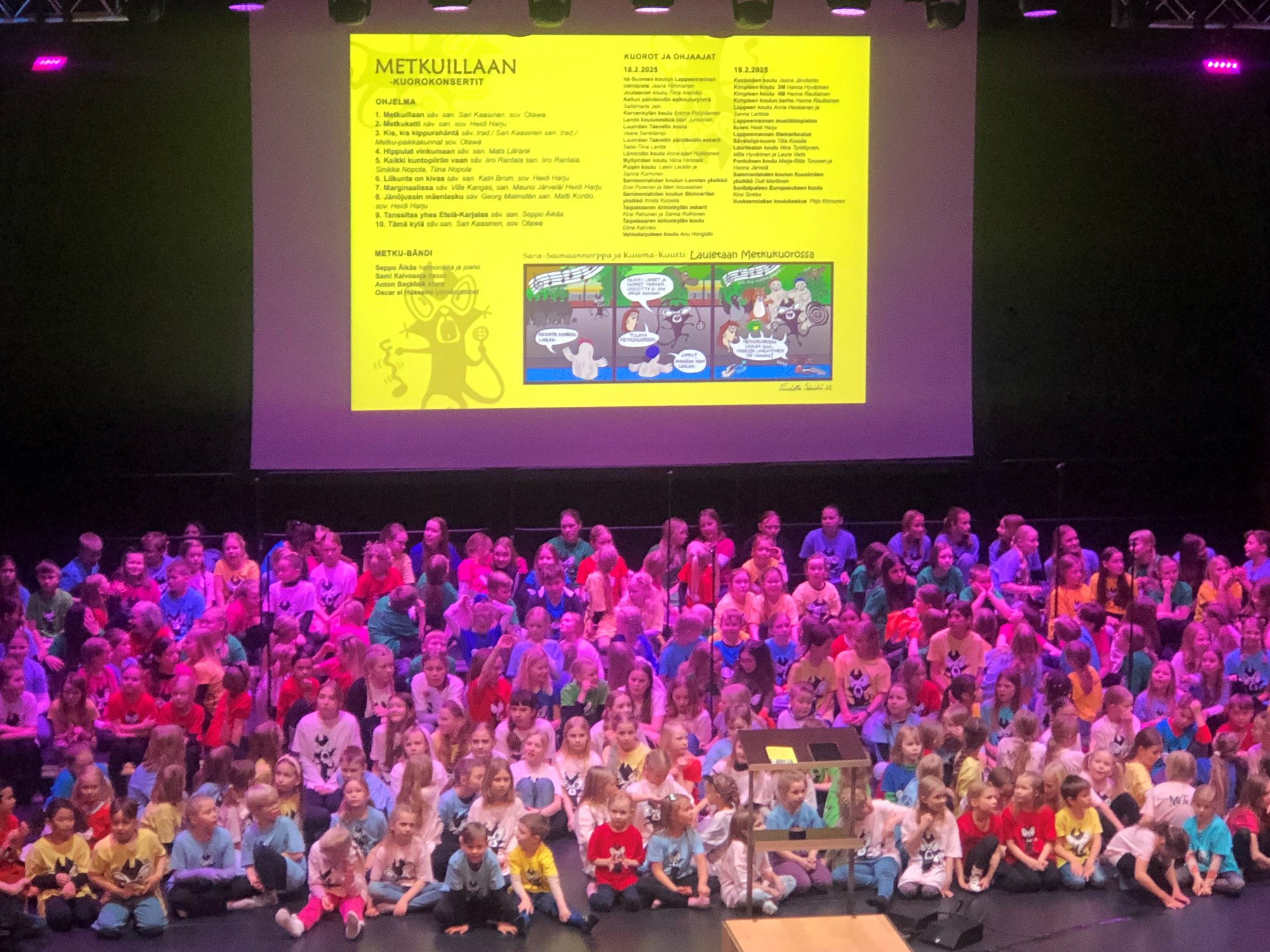 Lasten suurkuoro kokoontuneena istumaan esiintymislavalle, taustalla heijastettuna keltaisella pohjalla Metkuillaan-kuorokonserttien ohjelma.