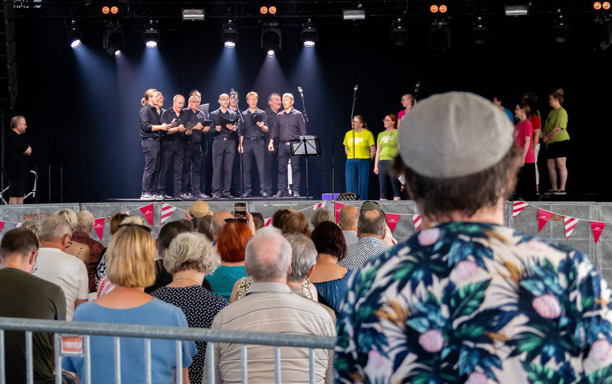 Yleisöä katsomassa Rannalla Saimaan -yhteislaulutapahtumaa, jossa kaksi kuoroa laulaa vastakkain lavalla.