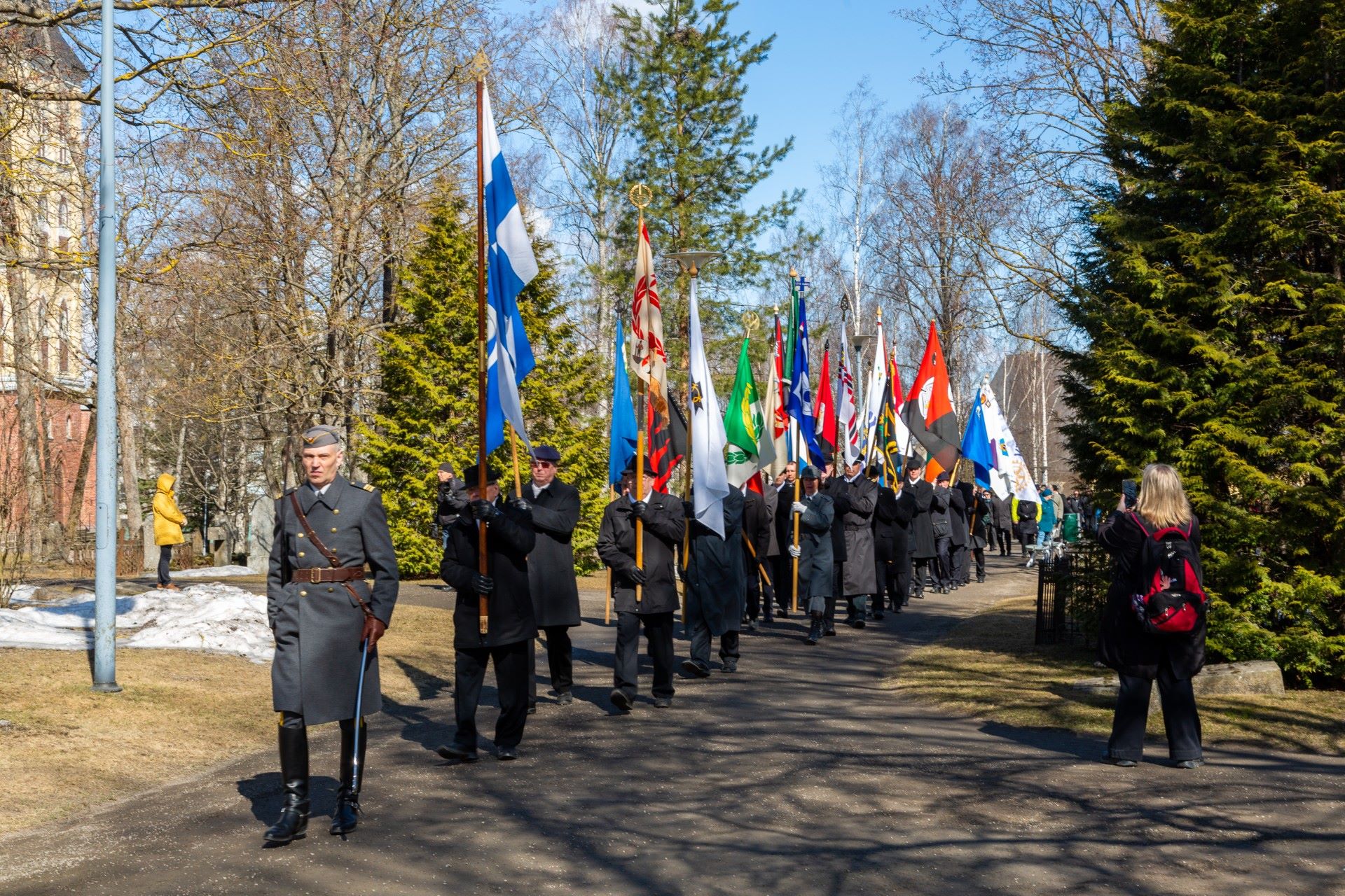 Sotilasasuun pukeutunut henkilö johtaa lippuja kantavia henkilöitä Lappeenrannan kirkkopuistossa.