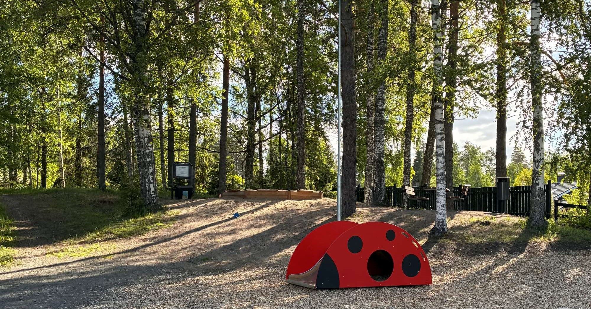 Leikkipaikka vihreässä puisessa maastossa. Punainen leikkilaite, kauempana hiekkalaatikko ja penkkejä.