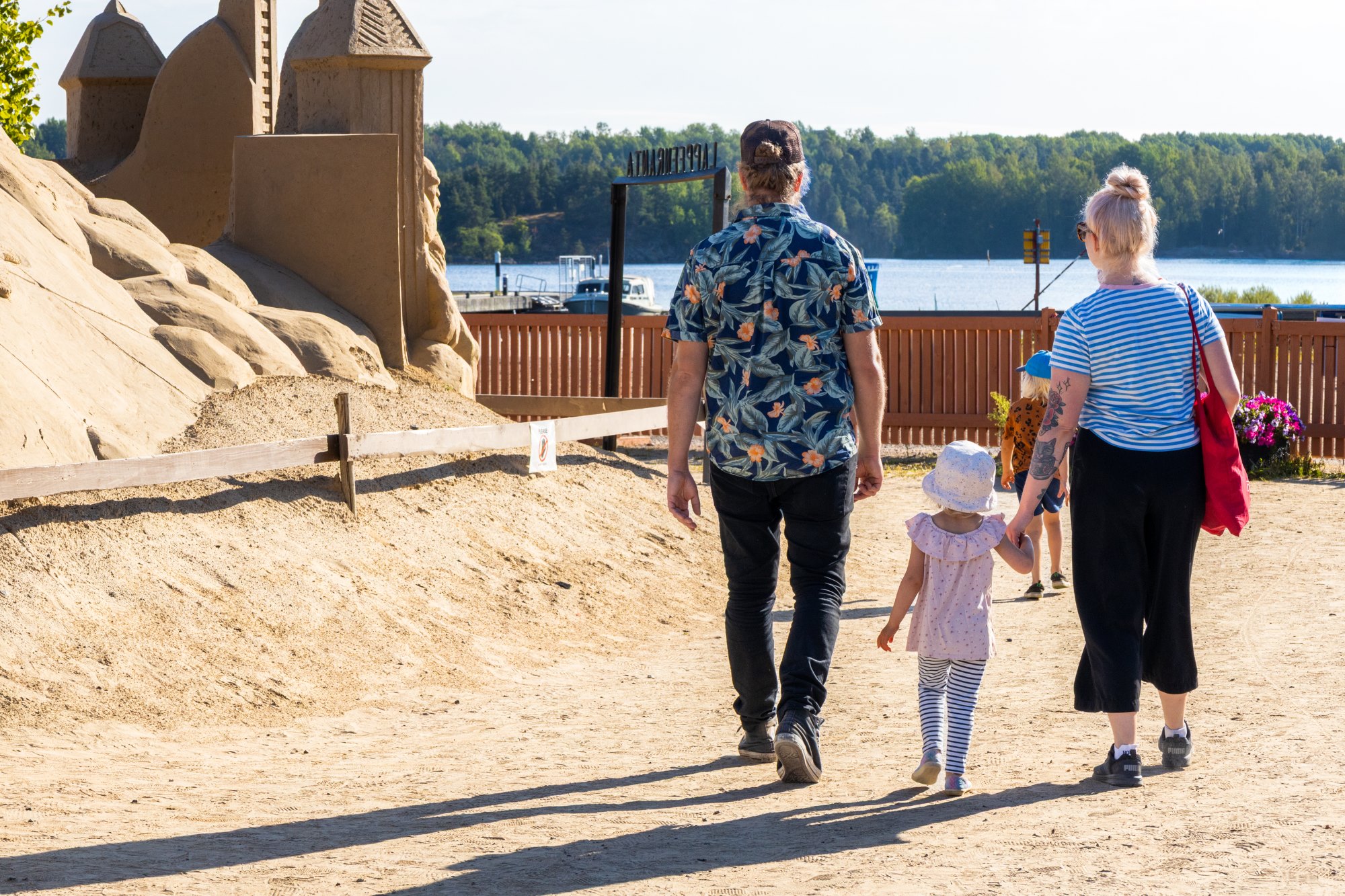 Kaksi aikuista ja kaksi pientä lasta kävelemässä hiekkalinnalla, taustalla Saimaa.