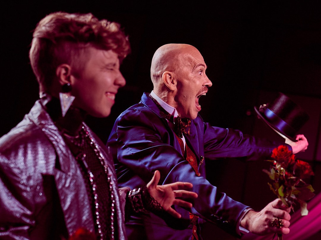 Kaksi juhlavasti pukeutunutta henkilöä seisoo tummaa tausta vasten. Molempien ilme on riemastunut ja heillä on käsissään ruusuja.